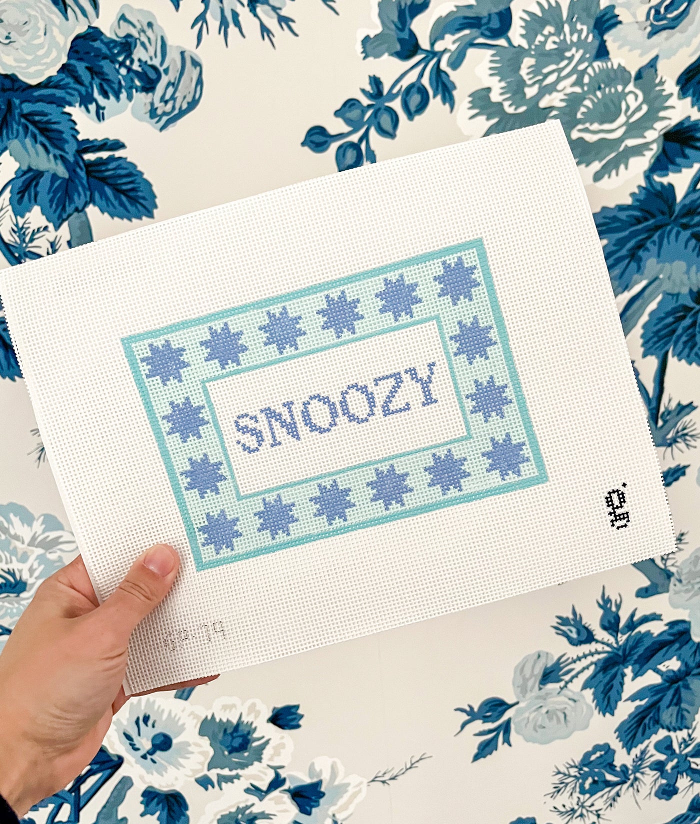 White needlepoint canvas with a aqua rectangular border with 6 pointed periwinkle stars surrounds a white center with the word "SNOOZY" in peri winkle text