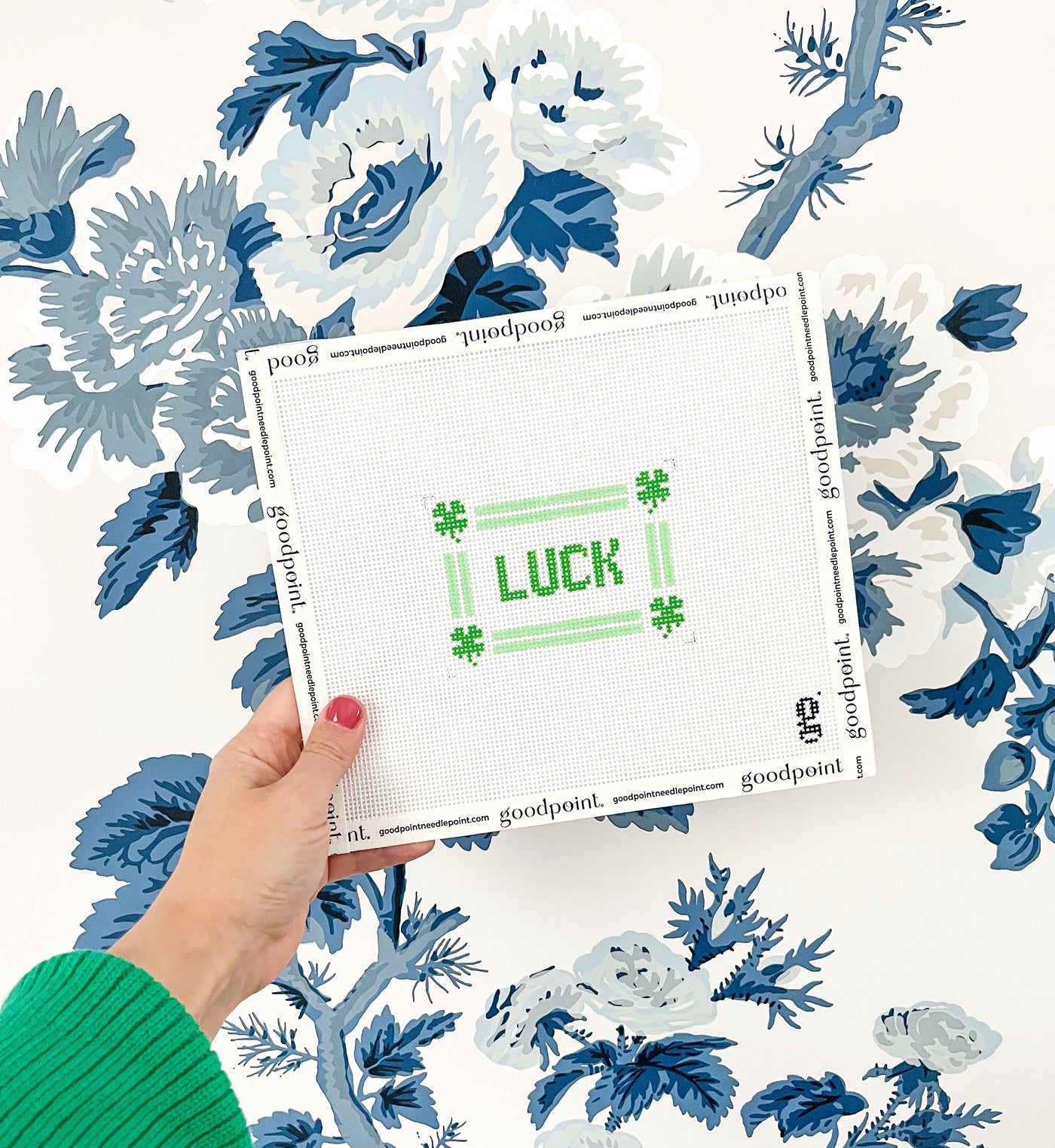 Hand holds up a white needlepoint canvas with the word LUCK in green text at center, surrounded by a mint green striped border and 4 leaf clovers at each corner