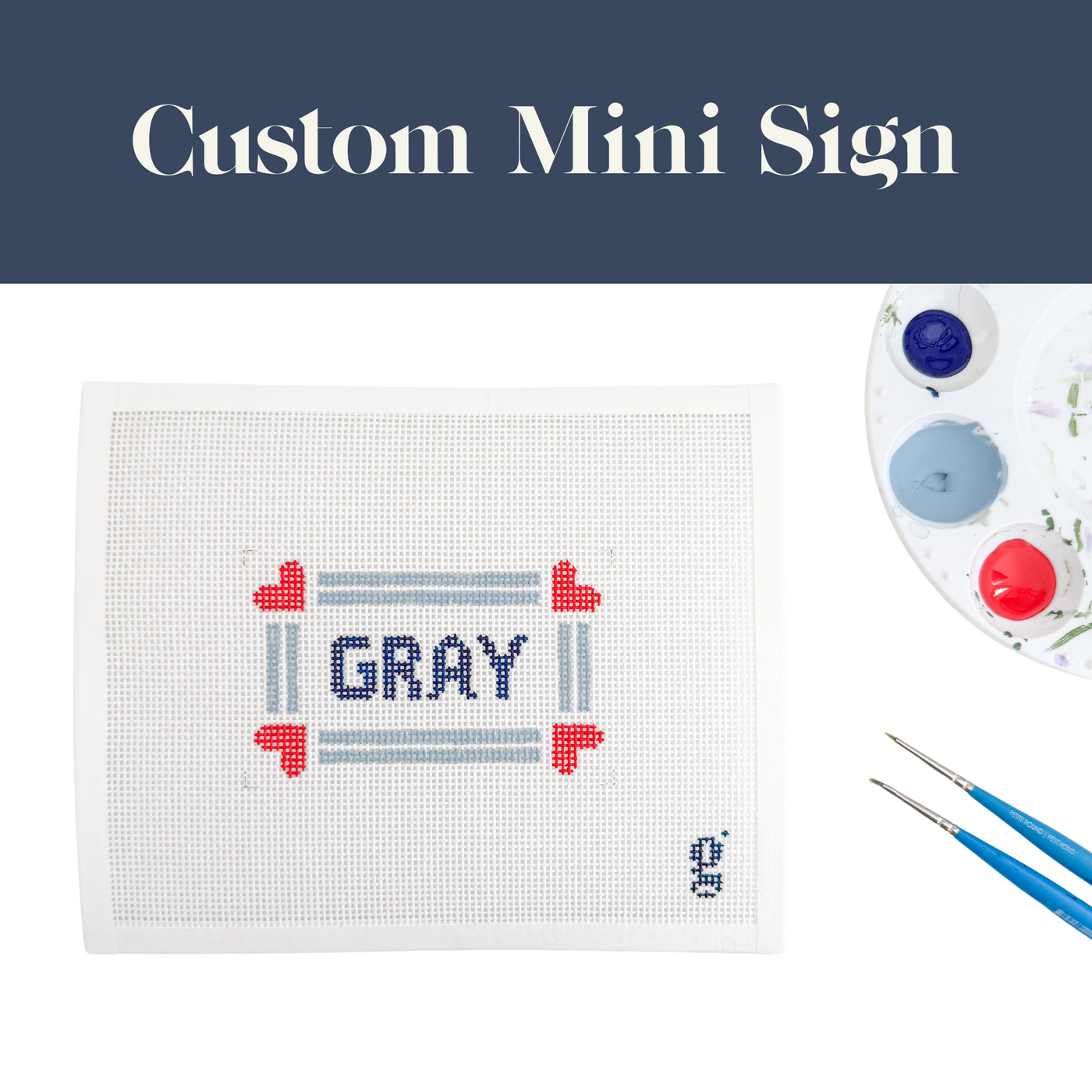 Paint palette and paint brushes sit to the side of a white needlepoint canvas with the name "GRAY" at center surrounded by slate blue striped border with red hearts at corners