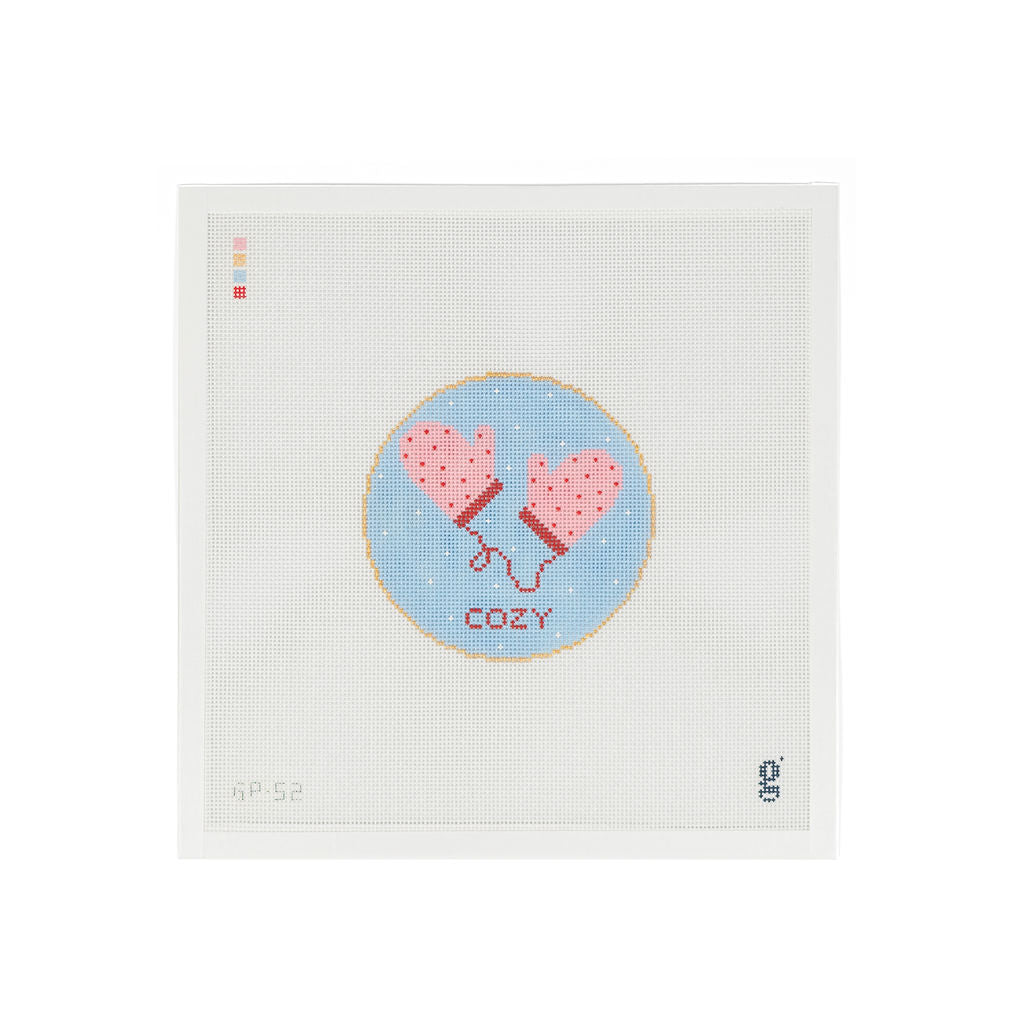 White needlepoint canvas with light blue circle at center. Inside the circle is a pair of pink mittens with red polka dots connected with a string. The word "COZY" is below in red text.