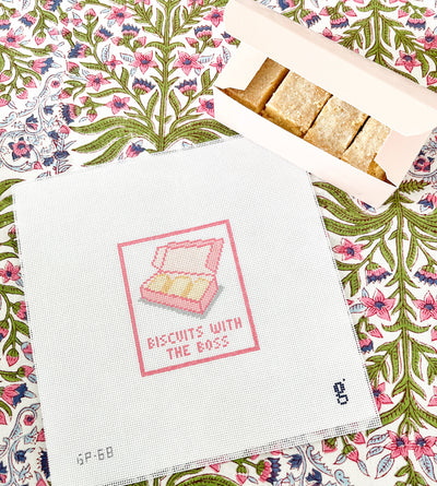 Needlepoint canvas with a pink box of shortbread and the words BISCUITS WITH THE BOSS sits on a floral tablecloth next to a pink box of shortbread