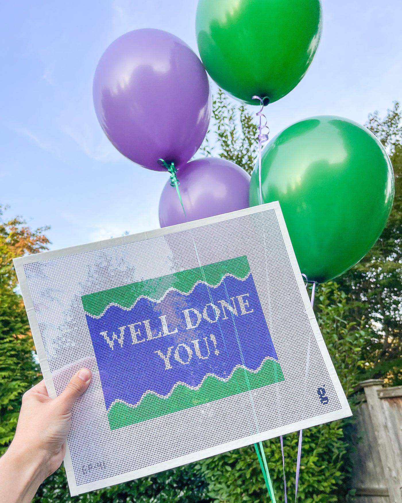 Purplish/blue & green needlepoint canvas with the words WELL DONE YOU in white font is held up in front of 4 balloons. 