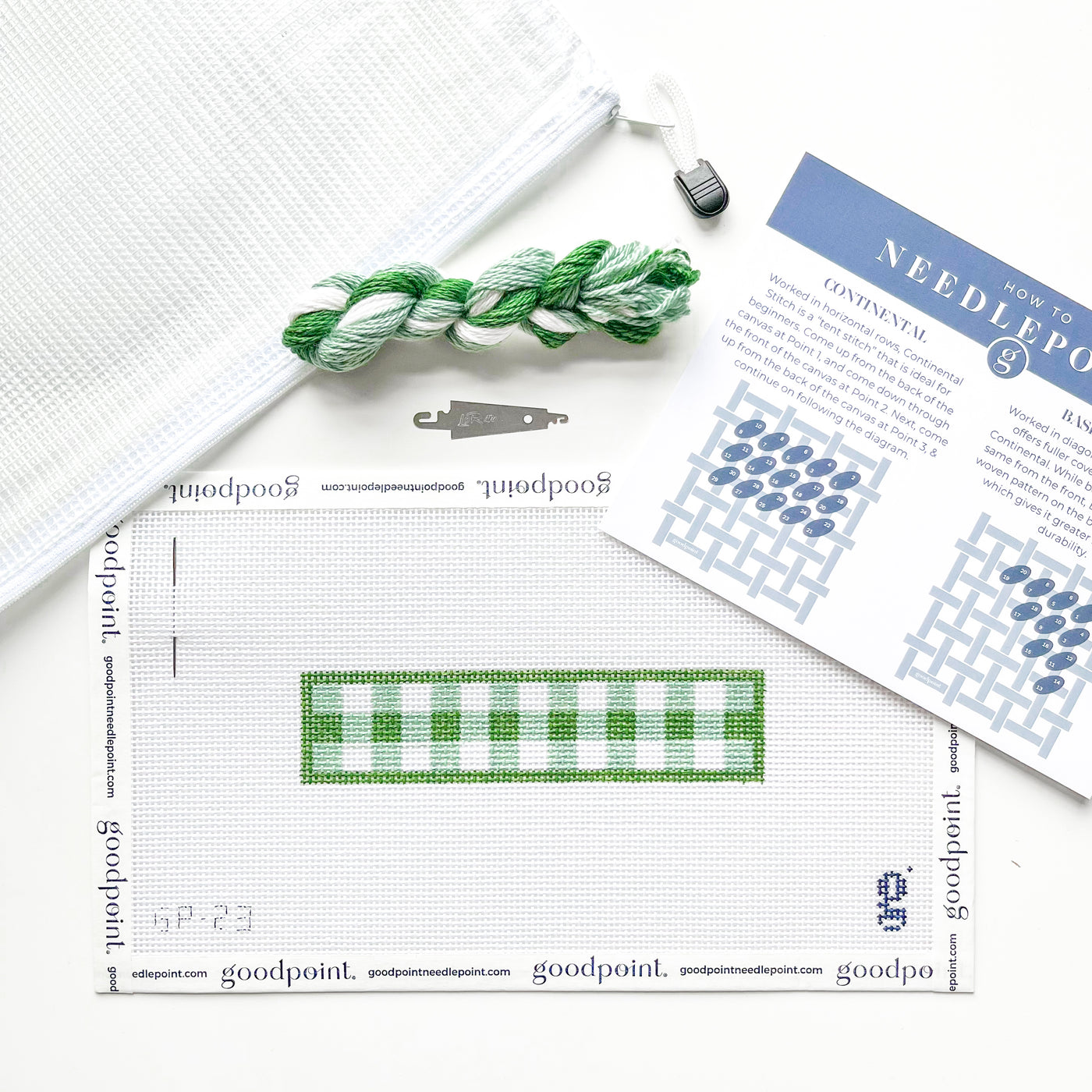 Green gingham key fob needlepoint canvas sits on white surface surrounded by needlepoint instructions, a needle threader, a bundle of threads and a white project bag